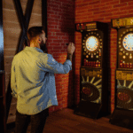 Players throwing darts at a board with numbered sections and a bullseye, surrounded by spectators and a scoreboard